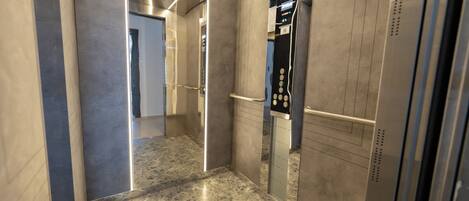 An elevator interior with metallic walls, a marble floor, and a control panel with buttons and a digital display.
