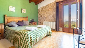 A bedroom with a wooden ceiling, a stone wall, a bed with a green and white checkered bedspread, and a view through a wooden door to the outside. - La Rosa de los Vientos – Charming Stay with Sea Views for Peaceful Days (La Serratella)