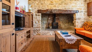 A stone-walled room with a television, wooden furniture, a fireplace, and an orange sofa. - La Rosa de los Vientos – Charming Stay with Sea Views for Peaceful Days (La Serratella)
