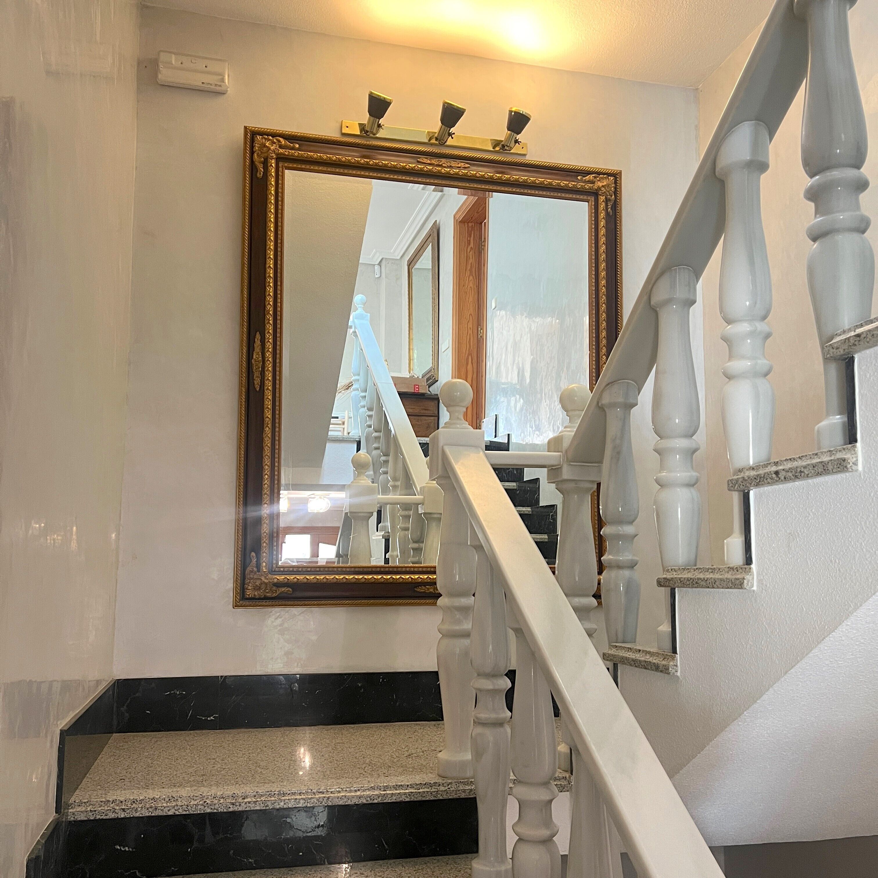 A staircase with white balusters and a black and white marble floor. A large mirror with a gold frame is mounted on the wall above the stairs.