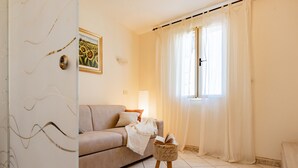 A living room with a sofa, a small table with a book, a lamp, and a window with curtains. - Apartment 'Anita' with Sea View, Private Terrace and Wi-Fi (Capoliveri)