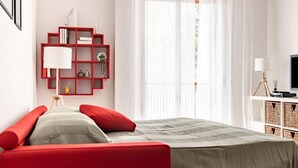 A modern bedroom with a red sofa, a bed with a grey bedspread, a red bookshelf, and a white desk with a lamp.