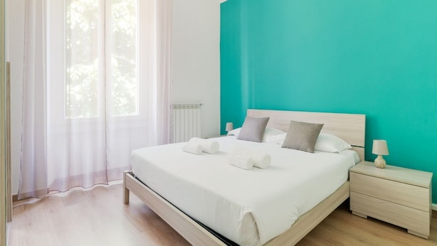 Een slaapkamer met een bed, nachtkastje, raam met dunne gordijnen en een wand in een turquoise kleur.
