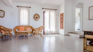 Een ruime woonkamer met een zithoek, twee ramen met gordijnen, een wanddecoratie en een trap naar een ander deel van het huis.