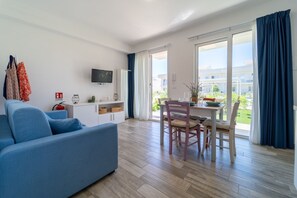 Ein modernes Wohnzimmer mit einer Couch, einem Esstisch, Stühlen, einem Flachbildfernseher und einer Glastür zum Balkon.