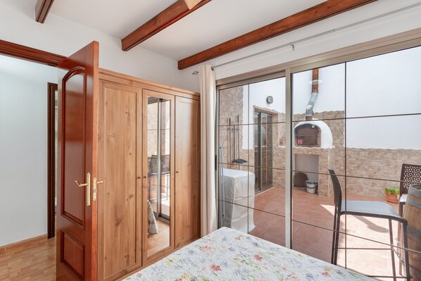 A room with a bed, wooden wardrobe, sliding glass door to an outdoor patio with a table and chairs, and a stone wall.