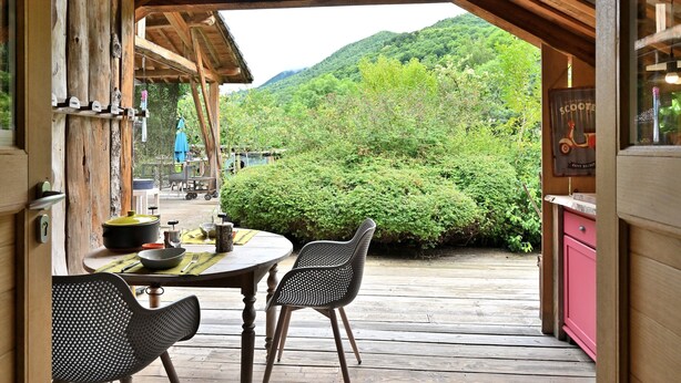 Eine Holzterrasse mit Tisch und Stühlen, Blick auf grüne Hügel und ein rustikales Blockhaus-Interieur.