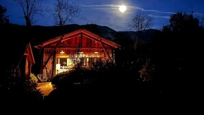Eine Holzhütte mit geneigtem Dach, umgeben von Bäumen, unter einem Nachthimmel mit sichtbarischem Mond.