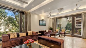 A spacious living room with a large sofa set, a coffee table, and a view of the outdoors through large windows. - Geet by Akama Homes (New Delhi)