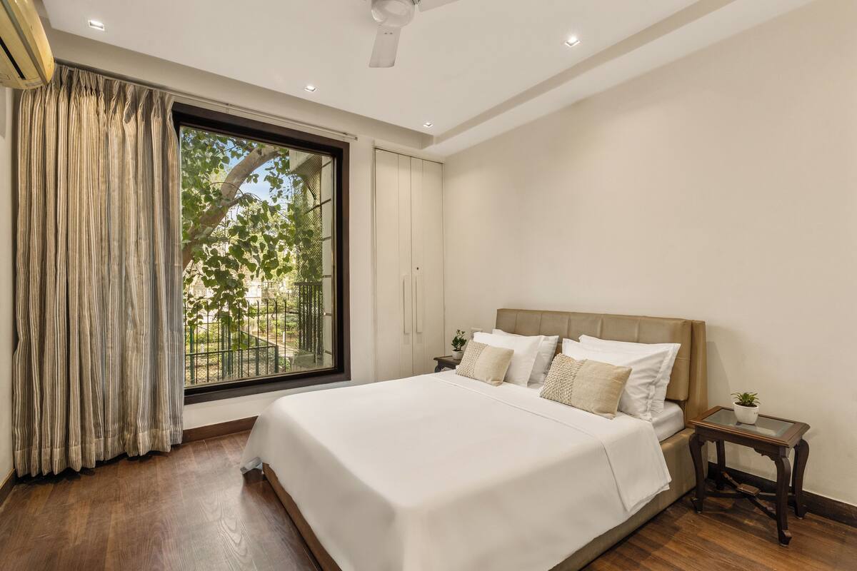 A bedroom with a large bed, a nightstand, a window with curtains, and a view of greenery outside.