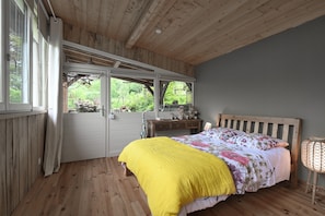 Un dormitorio con techo de madera, una cama con colcha floral, una mesita de noche y una ventana con vista a follaje.