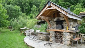 Une cuisine extérieure en pierre, dotée d’une cheminée, entourée d’un mur de pierre et d’une zone herbeuse.