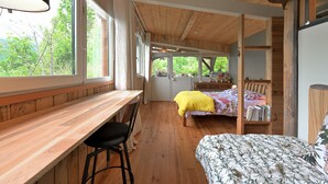 Une pièce avec un bureau en bois, un lit recouvert d’une couvre-lit à motifs floraux et une vue sur la verdure à l’extérieur.