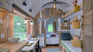 A rustic cabin interior with a wooden dining table, a bed with a green headboard, a hanging basket, and a window with a view of greenery. - Tiny Home 'Roulotte Appoline' with Mountain View, Private Terrace and Garden (Arrien-en-Bethmale)
