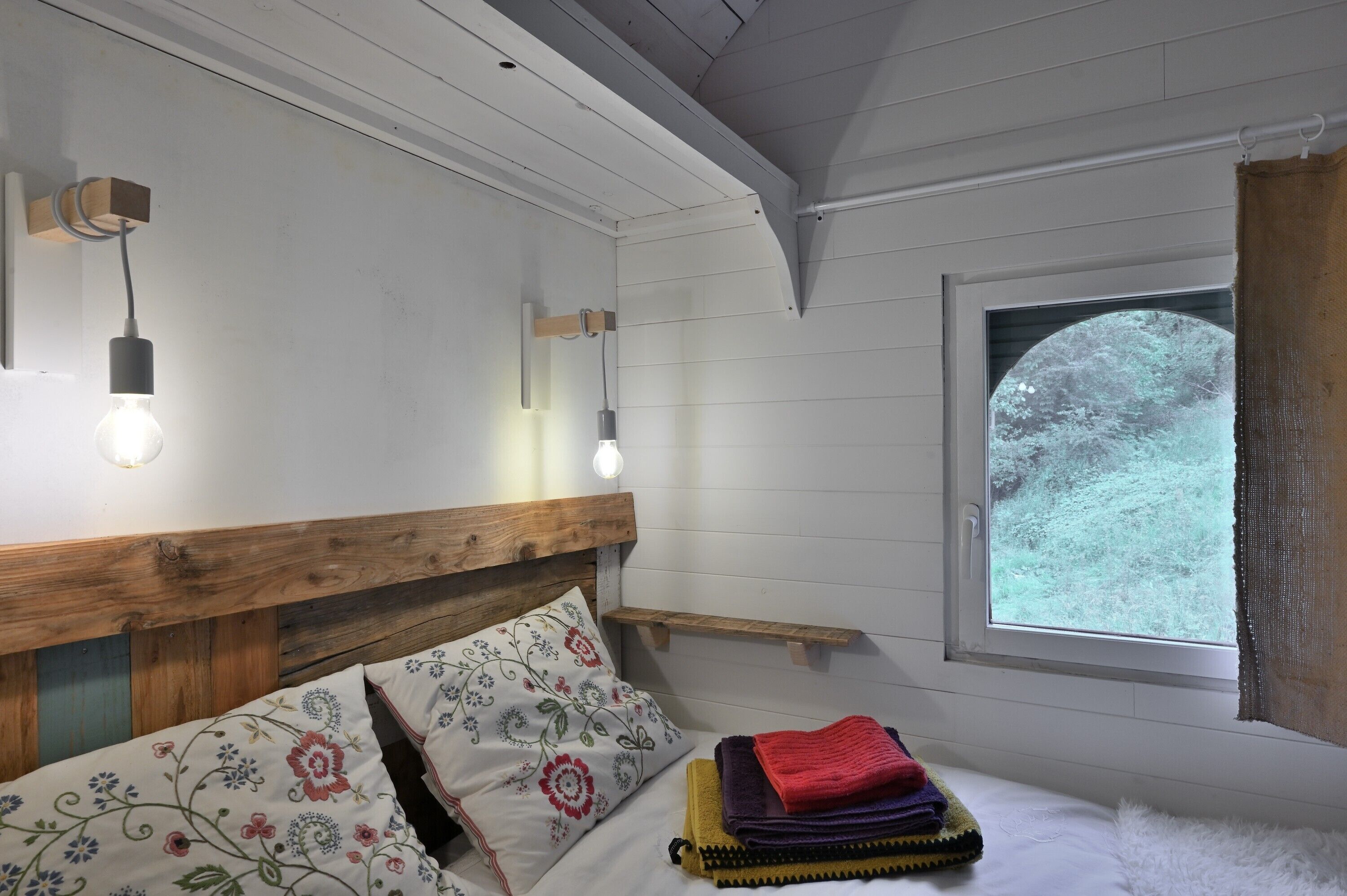 Une chambre à coucher avec une tête de lit en bois, un lit agrémenté d’oreillers à motifs floraux, une table de chevet, une fenêtre donnant sur des arbres et deux lampes fixées au mur.