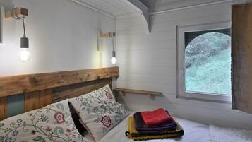 Une chambre à coucher avec une tête de lit en bois, un lit agrémenté d’oreillers à motifs floraux, une table de chevet, une fenêtre donnant sur des arbres et deux lampes fixées au mur.