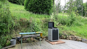 Een houten terras met een barbecue en een tafel, omgeven door groen en bomen.