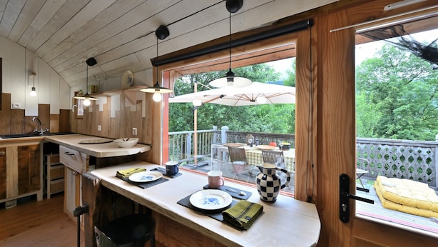 A wooden interior with a kitchen counter, dining area, and a view of the outdoors. - Tiny Home 'Roulotte Appoline' with Mountain View, Private Terrace and Garden (Arrien-en-Bethmale)