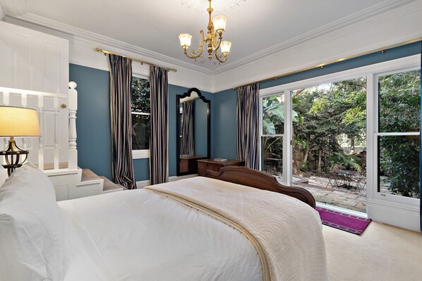 A bedroom with a large bed, a nightstand, a dresser, a mirror, and a chandelier. There is a window with curtains and a view of the outdoors.