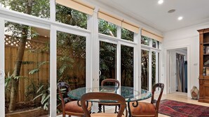 A dining area with a glass table and wooden chairs, large windows, and a view of a garden.
