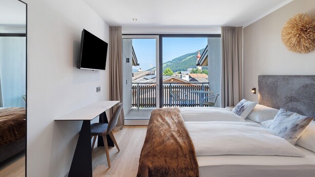 Een moderne hotelkamer met een bed, een bureau, een televisie en een balkon met uitzicht op gebouwen en bergen.