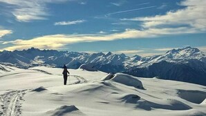Un paysage enneigé où l’on voit une personne marchant sur un sentier recouvert de neige, entourée de montagnes et sous un ciel dégagé.