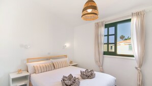 A bedroom with a bed, bedside tables, a hanging light fixture, a window with curtains, and a view of buildings outside.