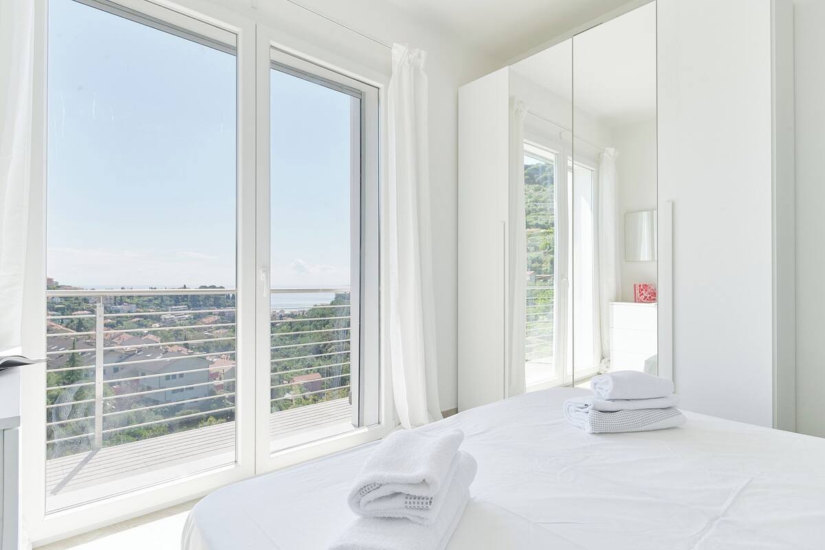 Ein Hotelzimmer mit großem Fenster, einem Bett mit weißer Bettwäsche und Blick auf die Stadtlandschaft.