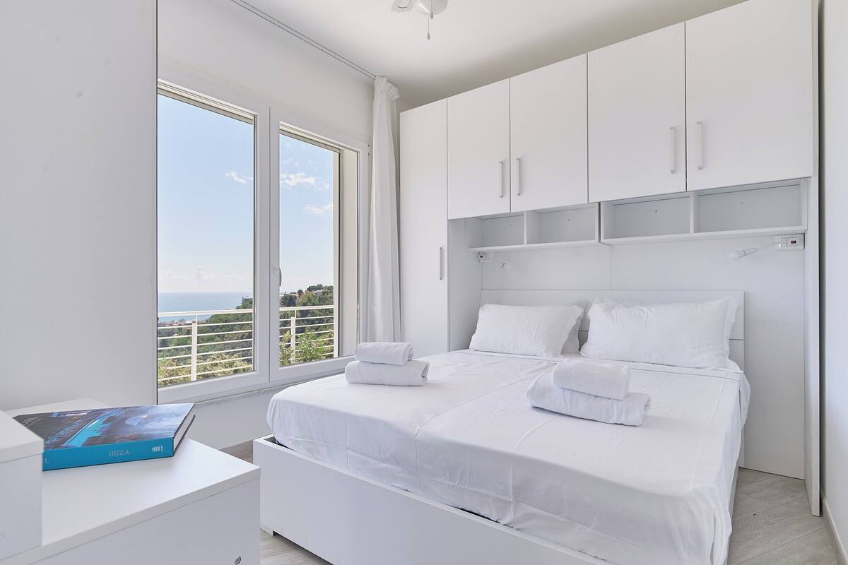Ein modernes Schlafzimmer mit einem großen Bett, weißer Bettwäsche und Meerblick durch ein großes Fenster.