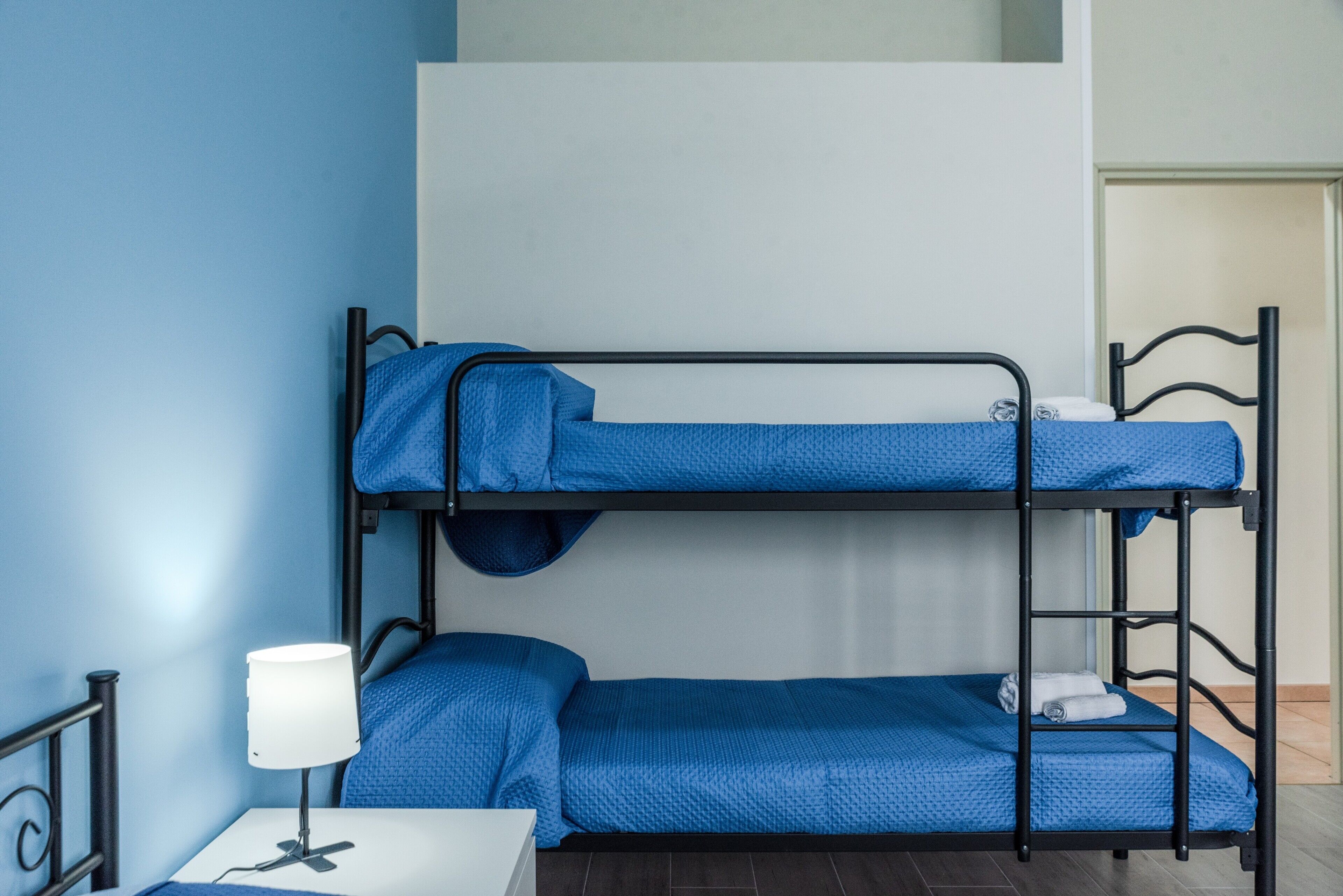 Een stapelbed met blauw beddengoed, een nachtkastje met een lamp, en een wandplank.
