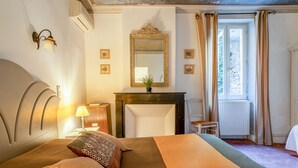 A bedroom with a bed, a nightstand, a lamp, a mirror, a fireplace, and a window with curtains. - "Orchidée" Room with Pool, Garden & Provençal Charm in Collias (Collias)