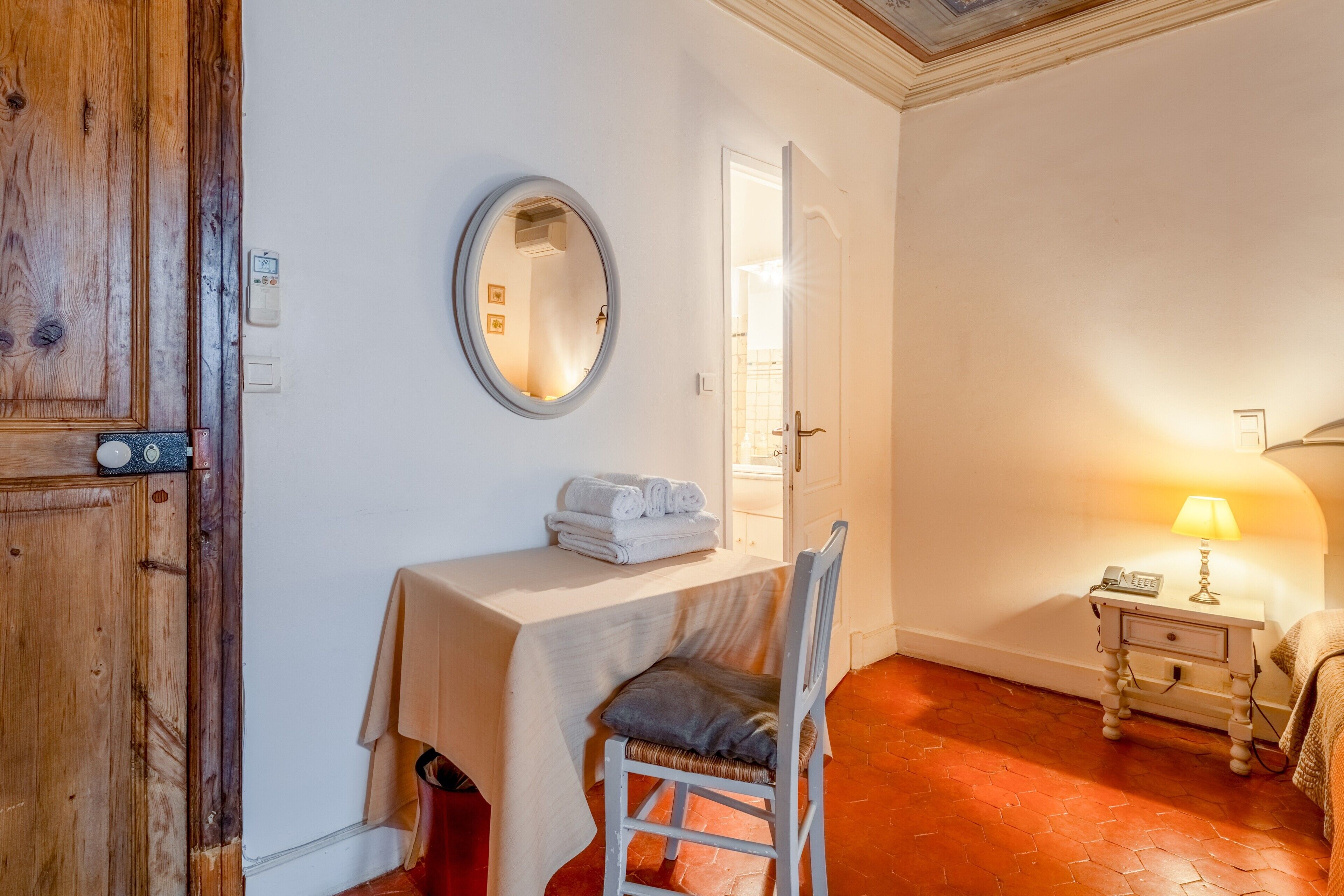 A room with a wooden door, a round mirror, a small table with a white tablecloth, a chair, a lamp, and a bed with a blanket.
