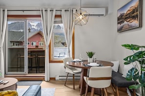 Een eetruimte met een tafel voor twee personen, een plant in pot en uitzicht op bergen door grote ramen.