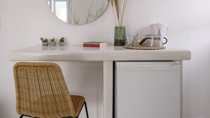 Un escritorio blanco con un espejo redondo, una silla de mimbre, un refrigerador pequeño y una cafetera.