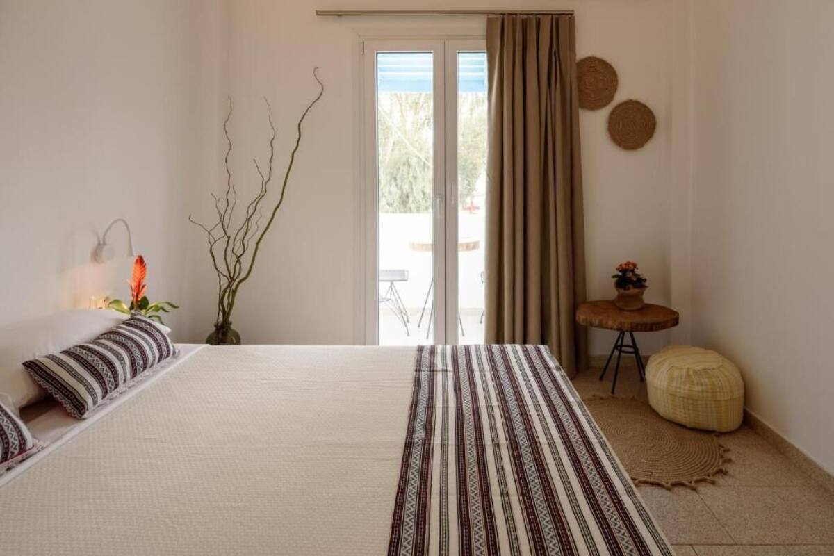 A bedroom with a large bed, a small table with a plant, a vase with flowers, and a view through a sliding door.