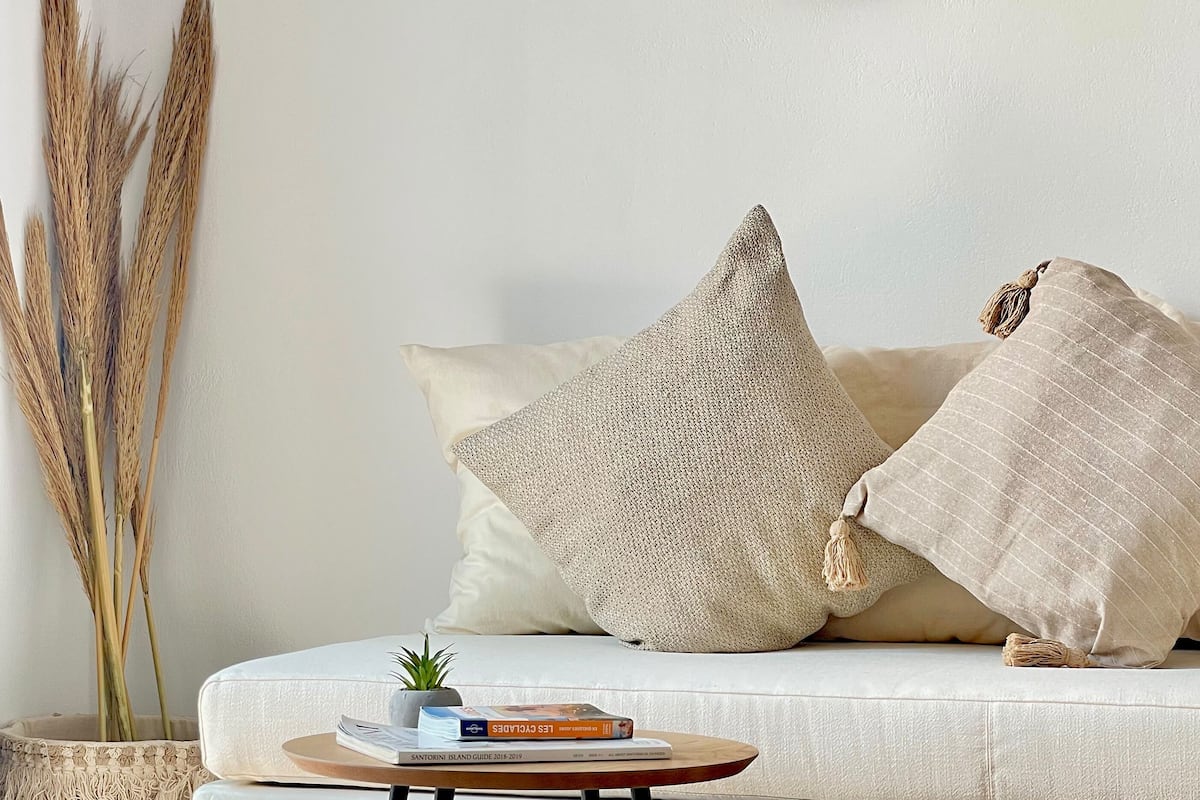A modern living room with a white sofa, two pillows, a round woven wall hanging, a small table with books, and a potted plant.