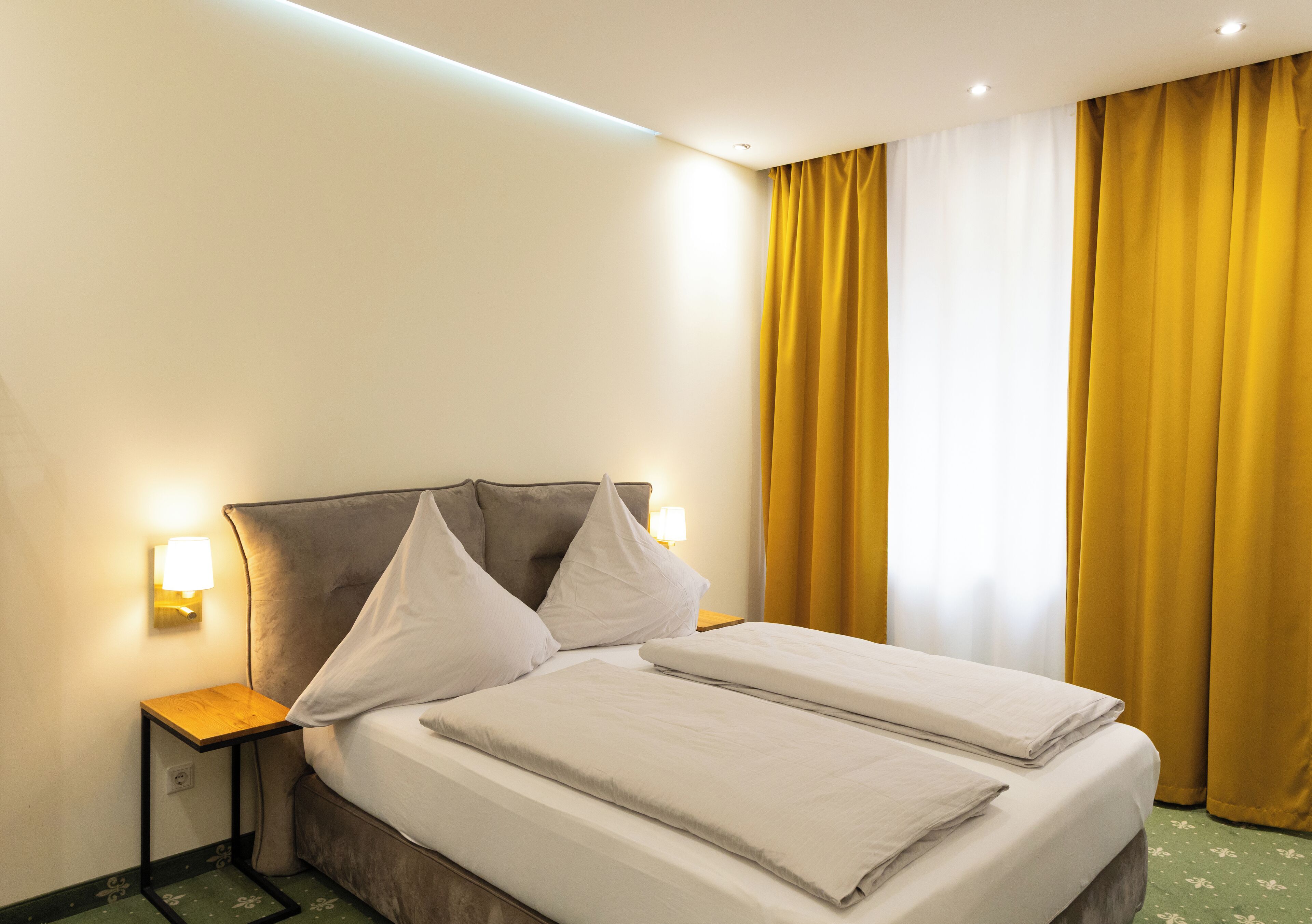A hotel room with a double bed, two bedside tables with lamps, a headboard, and yellow curtains.