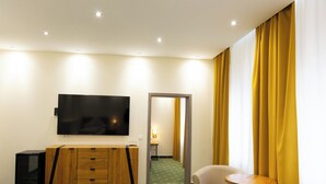 Quarto de hotel com televisão de tela plana, mobiliário de madeira, uma pequena mesa e cortinas amarelas.