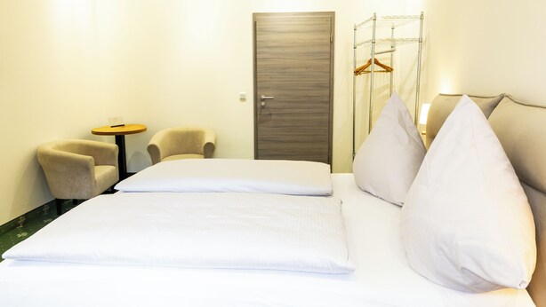 Quarto de hotel com uma cama, duas cadeiras, uma mesinha, um cabideiro e uma porta.