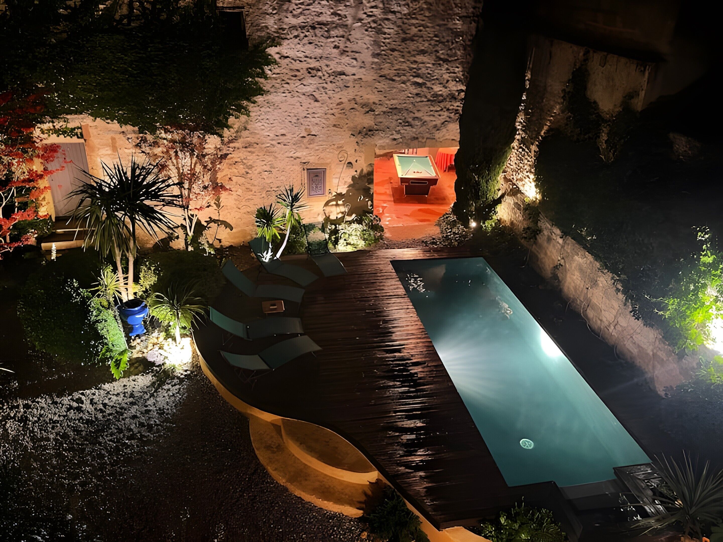 Une piscine dotée d’une terrasse en bois, entourée de plantes en pot et d’un mur de pierre.