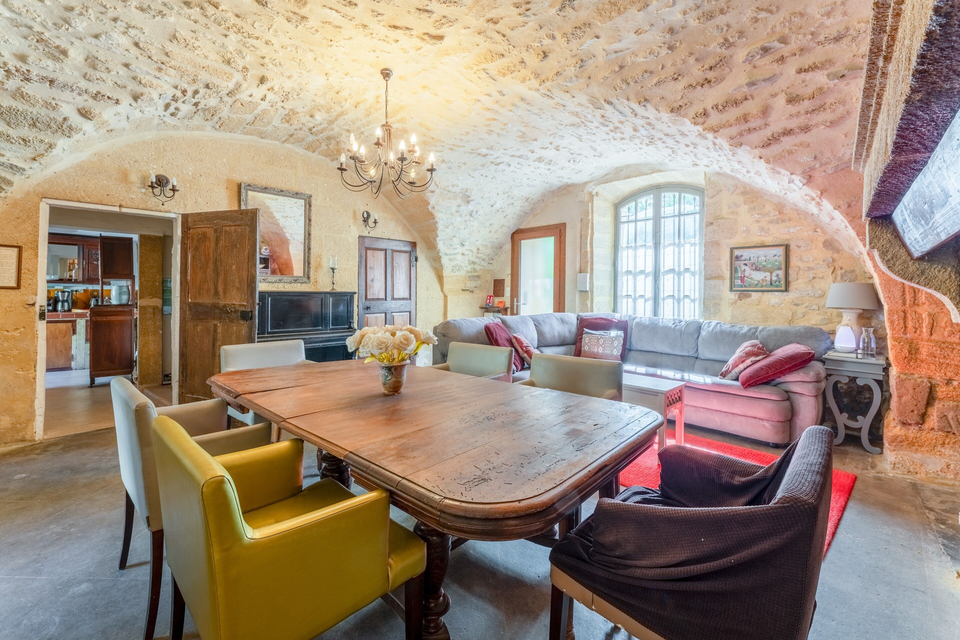 A spacious dining area with a wooden table and yellow chairs, a living room with a sofa and cushions, and a kitchen visible through an open doorway.