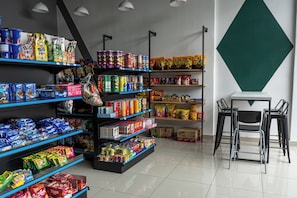 Een winkel met schappen vol snacks en drankjes, een zithoek met stoelen en een grote, groene, geometrisch vormgegeven wanddecoratie.