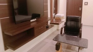 A modern room with a flat-screen TV mounted on a wooden cabinet, a small table with a chair, and a door with a mirror and towel stack.