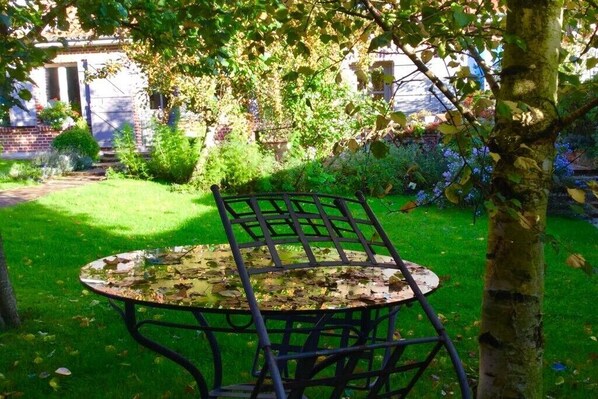 Un jardin avec une table ronde en métal et une chaise, entouré d’une végétation luxuriante et avec un bâtiment en arrière-plan.