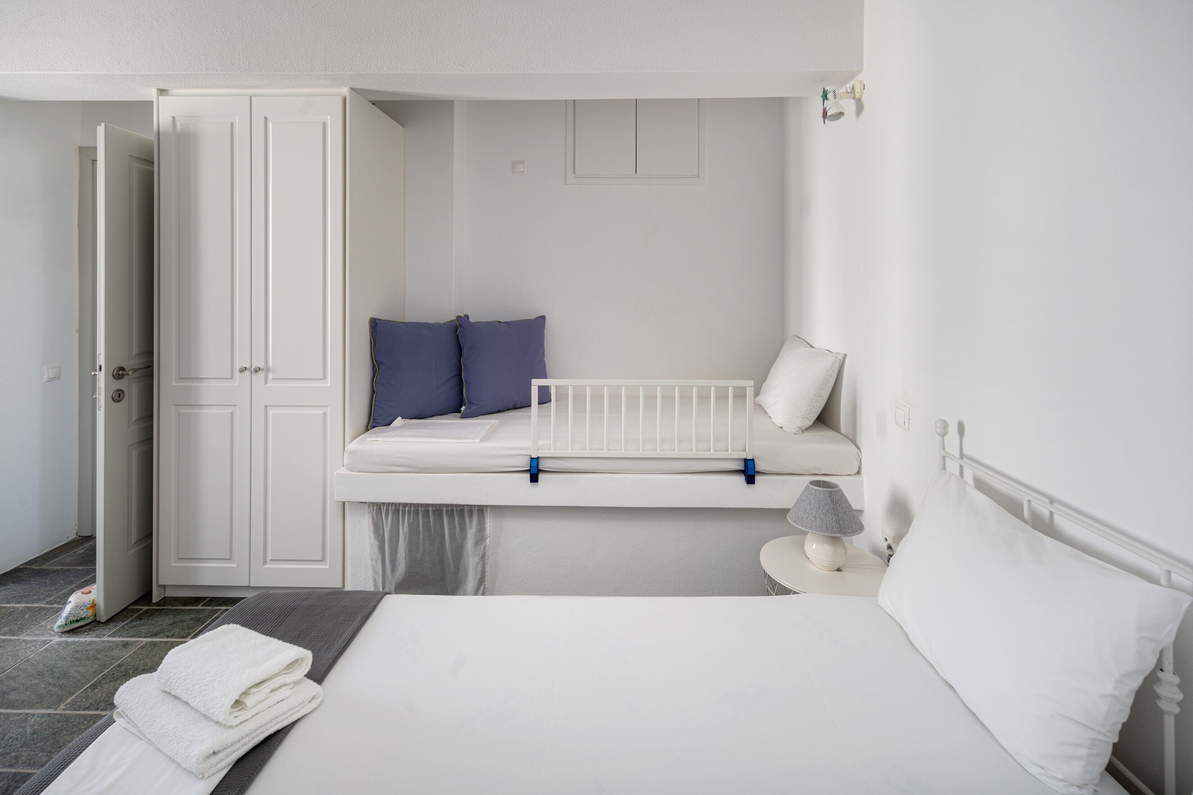 A bedroom with a bed, a nightstand, a window seat with pillows, a door, and a wardrobe.