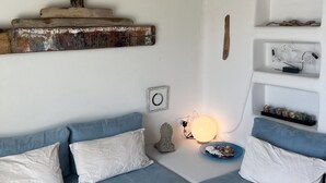 A modern living room with a blue sofa, white cushions, a wooden shelf with decorative items, a wall-mounted light, and a small round mirror.
