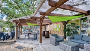 Un patio al aire libre con mesas y sillas de madera, sofás de mimbre y una zona de estar cubierta con toldos verdes.