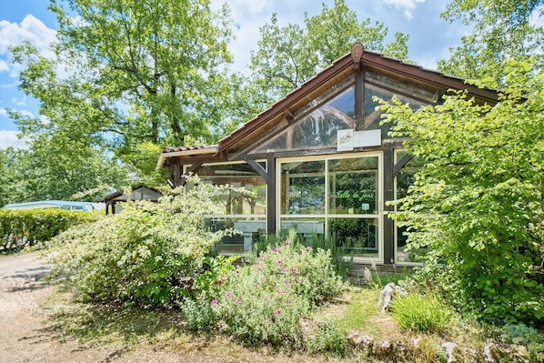 A glass-roofed building with a wooden structure, surrounded by lush greenery and a gravel path.