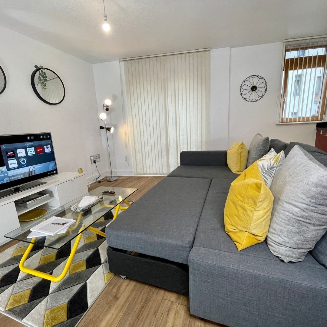 Un soggiorno moderno con un divano grigio, cuscini gialli e grigi, un tavolino da caffè in vetro e una TV a schermo piatto che mostra l'interfaccia di un servizio di streaming.