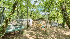 Un terrain de jeux avec des balançoires, un toboggan et un trampoline, le tout entouré d’arbres.
